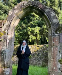 Eine Nonne steht unter einem alten Steinbogen in einer grünen Landschaft. Hinter ihr sind hohe Bäume und ein ruhiger, natürlicher Hintergrund zu sehen.
