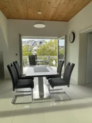 Ein helles Esszimmer mit einem weißen Tisch und schwarzen Stühlen. Große Fenster öffnen den Raum zur Terrasse mit Blick auf Bäume.