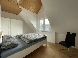 Ein helles Schlafzimmer mit einem Dachstuhl und Holzboden. Es gibt ein Doppelbett, einen Stuhl und einen Schrank.