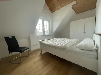 Ein einfaches Schlafzimmer mit einem Doppelbett und einem Stuhl. Die Wände sind hell und die Fenster bringen viel natürliches Licht herein.
