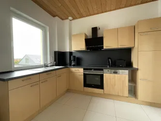 Eine moderne Küche mit hellen Holzmöbeln und einer schwarzen Arbeitsplatte. Ein großes Fenster sorgt für viel Tageslicht.