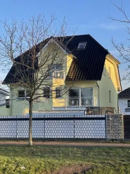 Ein modernes, gelbes Haus mit einem schwarz gedeckten Dach. Vor dem Haus steht ein Baum und ein Zaun aus Steinen.