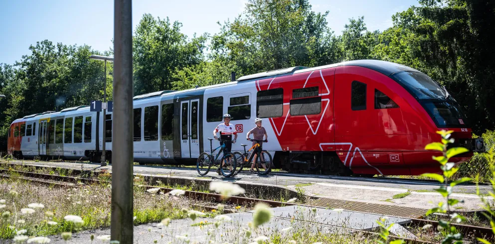 Zwei Fahrradfahrer schieben die Räder an einem Bahnsteig. Dahinter fährt ein roter Zug