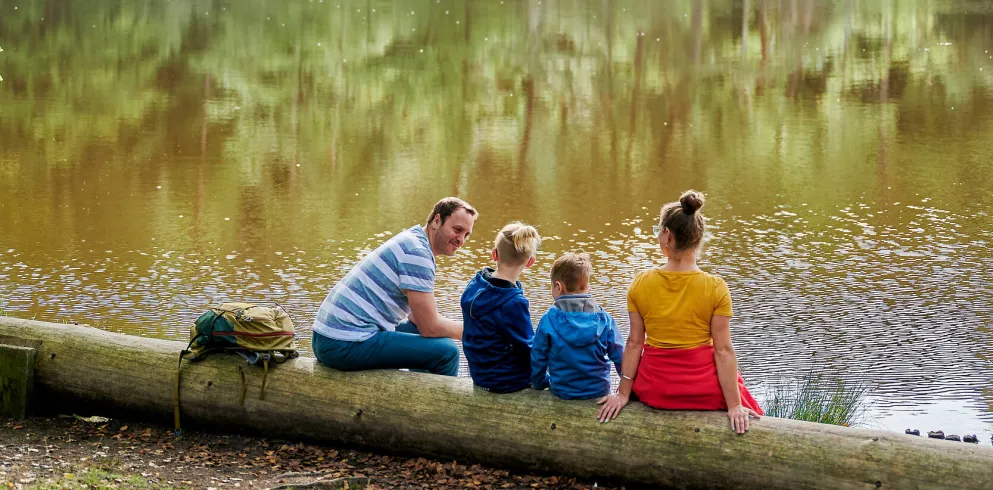 Familie sitzt auf Baumstamm direkt am Weiher