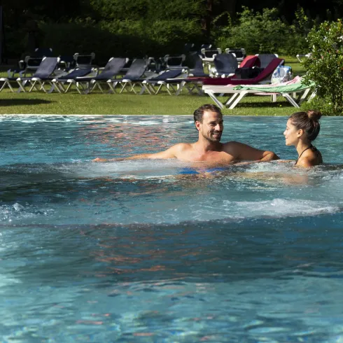 Ein Paar schwimmt in einem schönen Pool. Im Hintergrund sind Liegen und viel Grün zu sehen.