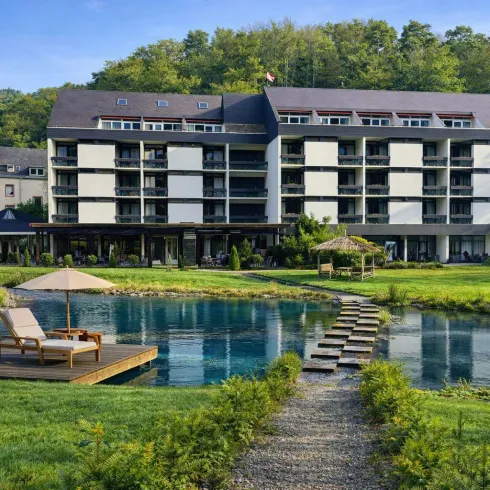 Ein modernes Hotelgebäude umgeben von üppigem Grün. Im Vordergrund befindet sich ein ruhiger Teich mit einer Holzterrasse und Liegestühlen.