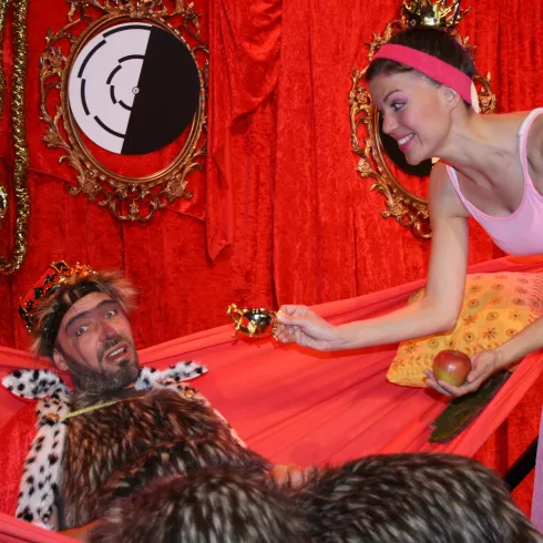 Eine humorvolle Theaterszene mit einem König in königlicher Kleidung, der auf einer roten Couch liegt. Eine junge Frau in einem pinken Kleid bietet ihm einen Apfel an.