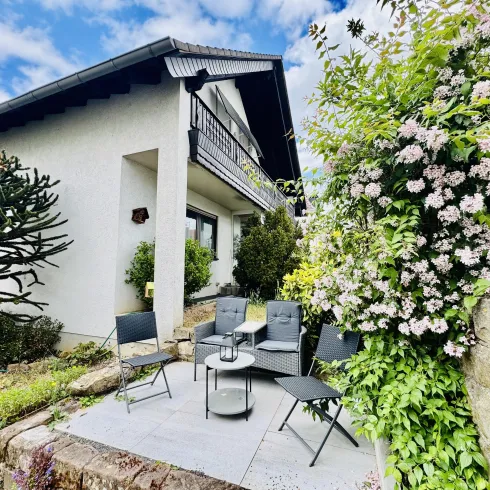 Ein gemütlicher Sitzbereich vor einem Haus, umgeben von grünen Pflanzen und blühenden Sträuchern. Die Terrasse bietet Platz für Entspannung im Freien.