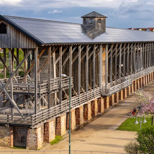 Ein historisches Holzgebäude mit einer offenen Struktur steht in einer malerischen Umgebung. Umgeben von blühenden Bäumen und einer weitläufigen Grünfläche.