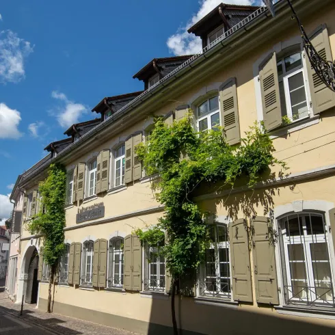 Ein historisches Gebäude mit traditionellen Fensterläden und einer begrünten Fassade. Der Eingang ist durch einen großen Torbogen gekennzeichnet.