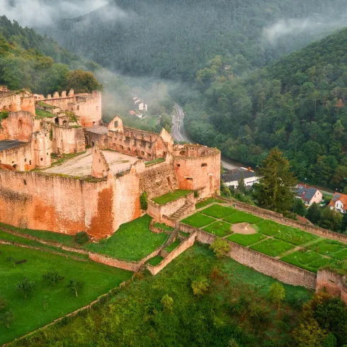 Eine alte Burruine umgeben von bewaldeten Hügeln und sanften Nebelschwaden. Der grüne Rasen und die teilweise erhaltenen Mauern verleihen der Szenerie noch mehr Charme.