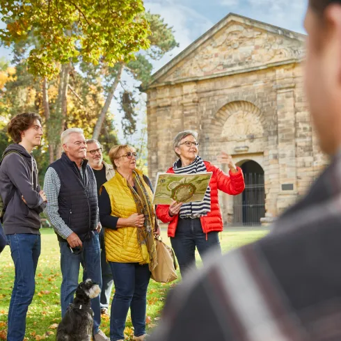 Eine Gruppe von Menschen steht im Freien und hört aufmerksam einer Frau zu, die eine Karte zeigt. Im Hintergrund ist ein historisches Gebäude und Bäume im Herbstlaub zu sehen.