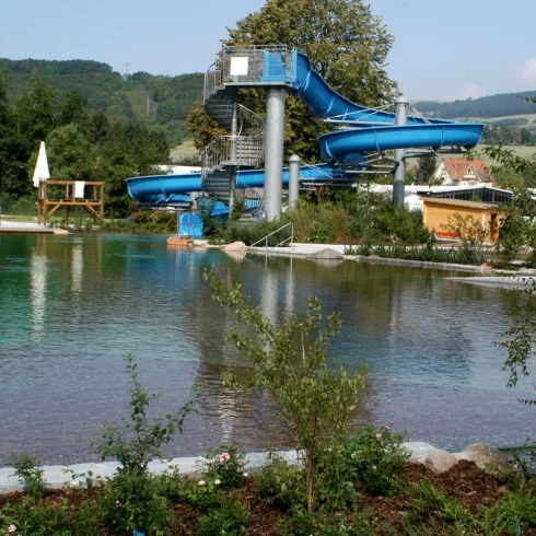 Ein Schwimmbad mit blauen Rutschen und klaren Wasserflächen. Im Hintergrund sind Bäume und eine grüne Landschaft zu sehen.
