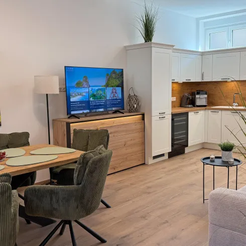 Ein modernes Wohnzimmer mit einer gemütlichen Couch und einem Esstisch. Die offene Küche ist hell und gut ausgestattet, mit einem großen Fernseher in der Ecke.