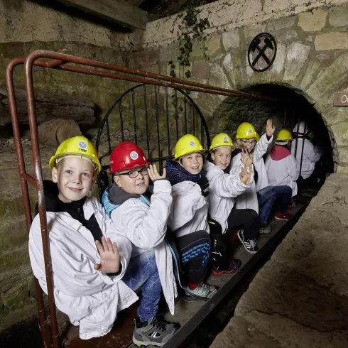 Eine Gruppe von Kindern sitzt in einem Tunnel und trägt Schutzhelme. Sie zeigen freundlich mit der Hand und tragen weiße Laborkittel.